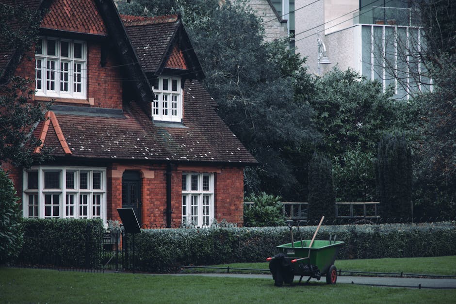 A red brick Victorian-style house with a steeply pitched roof and white-framed windows, surrounded by a well-maintained front garden. The garden features a neatly trimmed hedge along the front, lush g